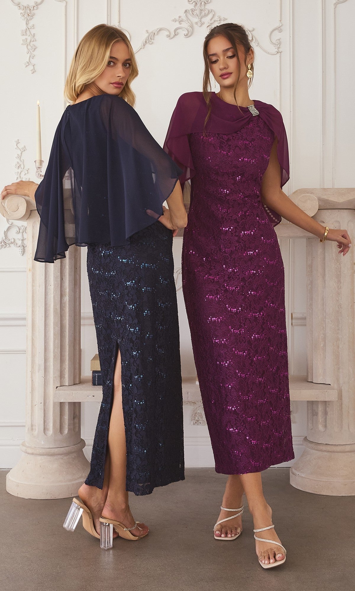 Two women in formal wear: one in the Amoris T906 long prom dress, a navy blue sequin-lace gown with sheer cape and side slit; the other wears a purple dress with matching chiffon cape overlay, both standing in an elegant room with classic columns.